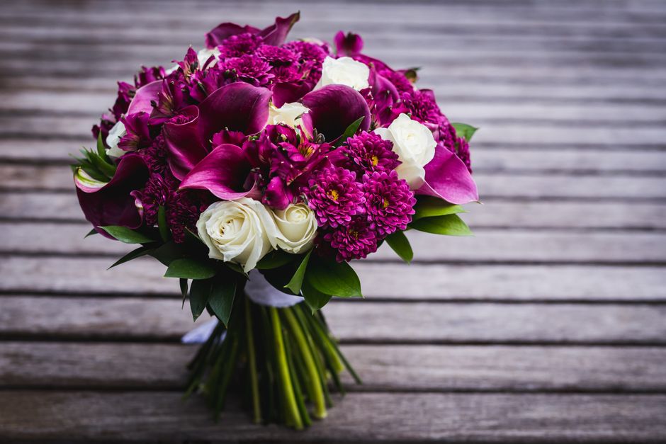 A bouquet of fresh floral arrangement featuring deep magenta calla lilies, bright purple dahlias, and clusters of white roses, all meticulously arranged with glossy green leaves. The bouquet is wrappe