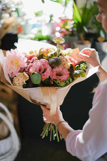 A person holding a large bouquet of fresh flowers, featuring pink peonies, cream-colored carnations, and small white and purple blooms, wrapped in beige and white paper with a satin ribbon. The arrang
