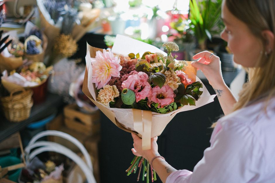 A woman with long blonde hair, wearing a light-colored top, holds a professionally arranged bouquet of fresh flowers, featuring pink dahlias, pale pink roses, purple and green foliage, and small white