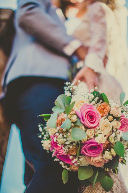 Close-up of a wedding bouquet arranged with pink and cream roses, accented with sprigs of baby's breath and eucalyptus leaves, held by a person in a wedding dress. In the background, a partially visib