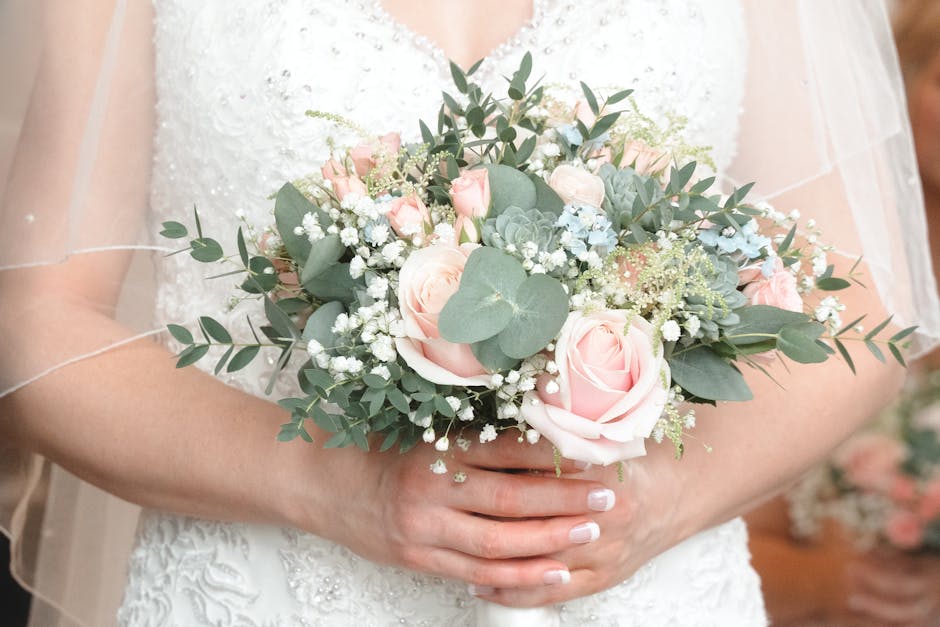 A bride holding a floral bouquet consisting of soft pink roses, white baby's breath, and green eucalyptus leaves, arranged in a rounded, lush style. The bouquet features a blend of pastel colors and f
